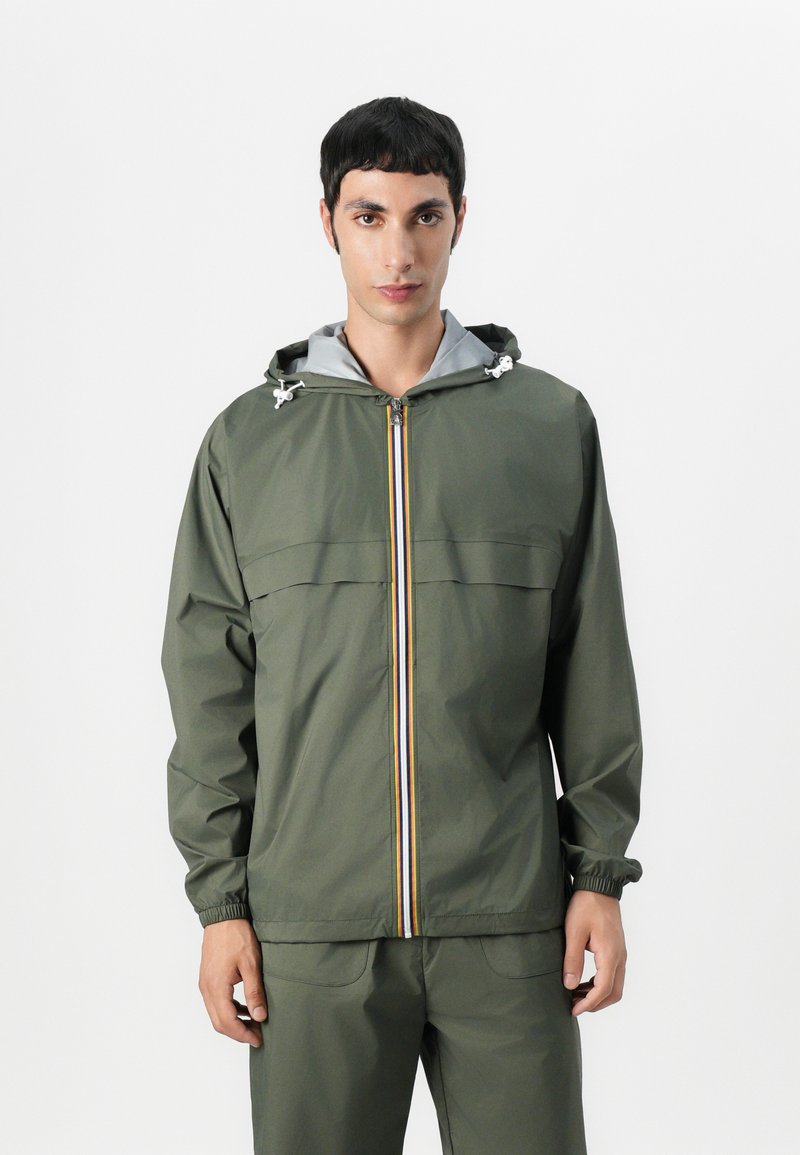Giacca leggera di colore verde oliva con zip frontale, fodera della cappuccio grigia, polsini elastici e una striscia verticale colorata al centro.