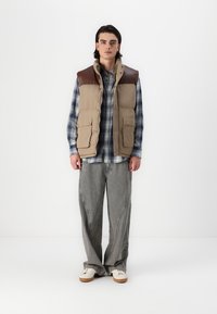 Beige Steppweste mit braunen Schulterakzenten, zwei vorderen Taschen, kombiniert mit einem blauen Karoshirt und hellgrauen gestreiften Hosen, abgerundet mit beigen Sneakers.