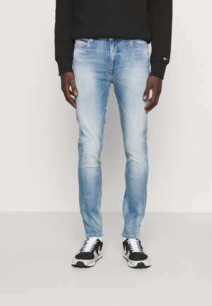 Mand iført lyseblå slim-fit jeans, sort sweatshirt og sorte og hvide sneakers, stående mod en ensfarvet baggrund.