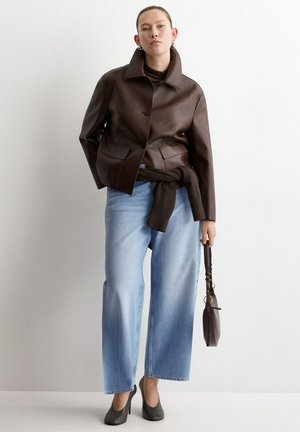 Mujer con chaqueta de cuero marrón oscuro, vaqueros de pierna ancha azul claro, tacones negros y sosteniendo un pequeño bolso marrón oscuro.