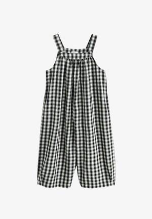 Mlada djevojka u crno-bijelim kariranim hlačama na tregere i bijeloj majici s dugim rukavima stoji na travi sa zatvorenim očima, rukama nježno na obrazima.