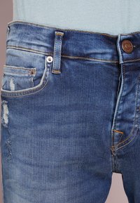 Modré džíny z denimu s vybledlým odstínem, obsahující opotřebované oblasti, žluté švy a kovové zapínání v pase.