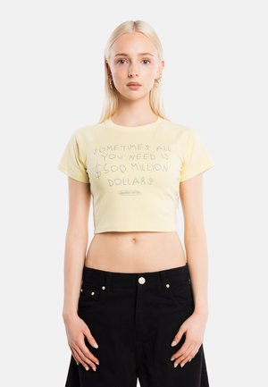 Joven con cabello largo y rubio lleva un top corto amarillo pálido con un texto sobre necesitar 500 millones de dólares y pantalones negros de talle alto.