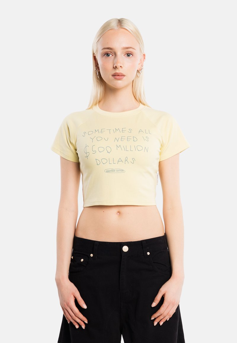 Jeune femme aux longs cheveux blonds porte un crop-top jaune pâle avec un texte parlant du besoin de 500 millions de dollars et un pantalon noir taille haute.