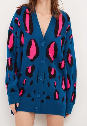 Femme portant un cardigan bleu surdimensionné avec un grand imprimé léopard rose et noir et des boutons bleus, montrée des épaules jusqu'à mi-cuisse.