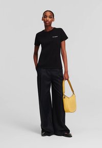 Sieviete, tērpusies melnā T-kreklā un platajās biksēs, tur dzeltenu rokassomu, stāvot pretī vienkāršam gaiši pelēkam fona.