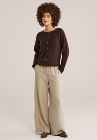 Sieviete stāv ar rokām kabatās, valkājot tumši brūnu ar pogām cieši pieguļošu džemperi, bēšas krāsas platas bikses un apavus ar nagliņām uz vienkārša fona.