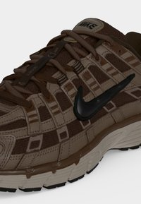 Καφέ και μπεζ αθλητικό παπούτσι Nike με διακοσμητικό επάνω μέρος, μαύρο λογότυπο Swoosh και καφέ κορδόνια σε λευκή σόλα και ουδέτερο φόντο.