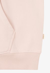 Tissu de sweatshirt rose clair avec un ourlet côtelé et une poche. Texture lisse avec des détails de couture visibles. Petite étiquette sur la couture latérale.