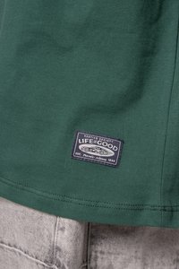 Green cotton t-shirt with a woven label featuring "Life is Good" and a vehicle graphic, sewn onto the bottom hem.