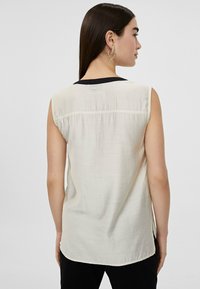 Blusa sin mangas en tejido blanco roto con un cuello negro; presenta una costura en la parte posterior y un botón decorativo en el escote. Textura suave y corte relajado.
