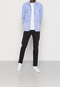 Ljusblå denimjacka med två bröstfickor, bärs över en vit T-shirt och svarta slim-fit jeans, kompletterad med vita sneakers.