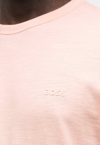 Κοντινό πλάνο ενός ατόμου που φοράει ένα ανοιχτό ροζ μπλουζάκι με στρογγυλή λαιμόκοψη και μικρό λογότυπο "BOSS" στο στήθος.