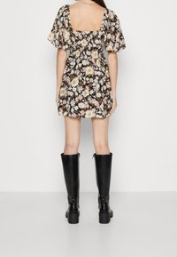 Florales Minikleid in Schwarz mit kurzen Puffärmeln, ausgestattet mit einem quadratischen Rückenausschnitt, tailliertem Bund und einem fließenden Rock. Kombiniert mit kniehohen schwarzen Stiefeln.