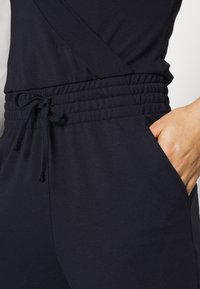Jogger en tissu noir avec une taille élastiquée, un cordon de serrage ajustable et une poche latérale. Texture lisse, design décontracté, coupe ajustée.