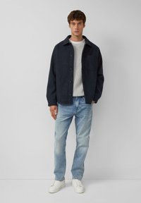 Marineblaues Hemdjacke mit Kragen, kombiniert mit einem hellgrauen gerippten Pullover, hellblauen Jeans und weißen Sneakers. Schlichtes, modernes Design.