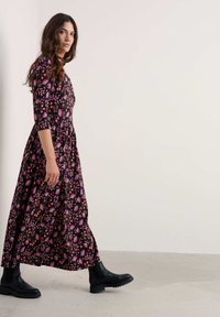 Duga crna haljina s ružičastim i crvenim cvjetnim uzorkom, rukavi do tri četvrtine, slojevita suknja, uparena s crnim čizmama do gležnja i teksturiranom površinom.