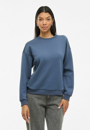 Sweatshirt bleu à encolure ronde et épaules tombantes. Tissu doux avec des poignets et un ourlet côtelés. Associé à un pantalon gris à rayures.