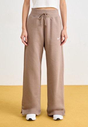 Person iført løstsiddende brune joggingbukser med snor i taljen og hvide sneakers på gul gulv.