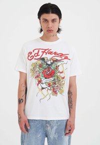 Camiseta blanca de algodón con gráficos inspirados en tatuajes en colores vivos y texto en cursiva roja con motivos florales. Corte relajado y mangas cortas.