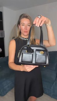 Fekete bőr kézitáska két első zsebbel, arany színű kiegészítőkkel és rövid fogantyúkkal. Az elején egy címke található, és lekerekített alakja van.