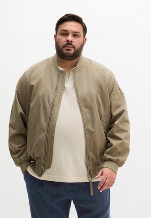 Mann mit Bart, der eine beige Bomberjacke mit Reißverschluss, ein cremefarbenes strukturiertes Hemd und marineblaue Hose trägt, steht vor weißem Hintergrund.