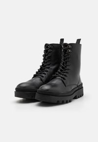 Botas de cuero negras hasta el tobillo con suela gruesa, parte delantera con cordones y ojales reforzados. Cuentan con una textura suave y una lengüeta para facilitar su puesta.