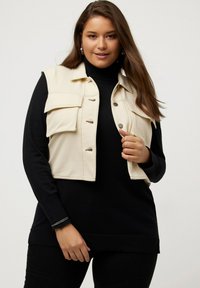 Gilet beige corto con quattro tasche, bottoni a scatto argentati, indossato sopra un dolcevita nero. Modello in posa con le mani sui fianchi, espressione rilassata.
