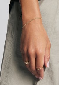 Złoty łańcuszek na nadgarstku, składający się z małych ogniw. Dłoń ozdobiona dwoma dodatkowymi złotymi pierścionkami, z których jeden ma okrągłe koraliki, a drugi wzór chevron.