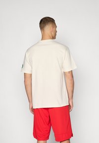 Kremfarget bomulls t-skjorte med korte ermer, avslappet passform, og en grønn logo på venstre erme. Stylet med røde shorts.
