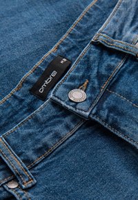 Jean en denim bleu foncé avec un bouton argenté, des détails de couture visibles, et une étiquette noire portant l'inscription « ombre » près de la ceinture.