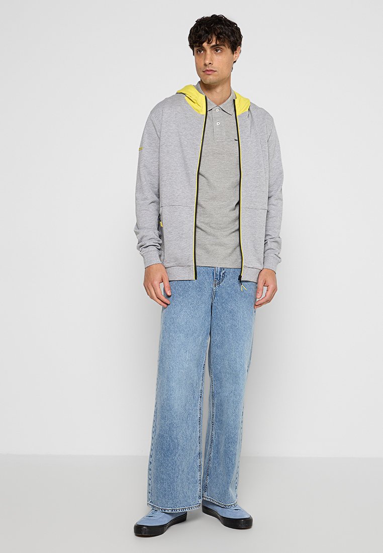 Sweat-shirt zippé gris clair avec des accents jaunes sur un t-shirt gris, associé à un jean bleu à jambes larges et des chaussures noires. Tissu lisse, coupe décontractée.