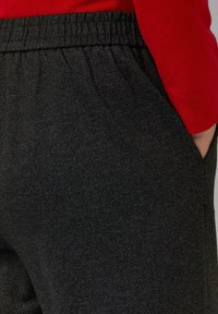 Dunkle Hose mit Fischgrätenmuster und elastischem Bund. Tasche auf der rechten Seite sichtbar, kombiniert mit einem roten Langarmshirt.