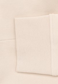 Lichtbeige sweatshirtstof met een gestructureerde afwerking. Heeft een grote, gestikte zak en zichtbare naaddetails, wat zorgt voor een schone, eenvoudige uitstraling.