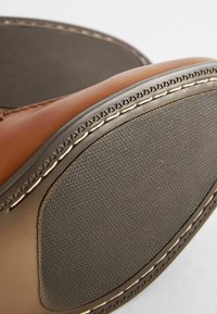 Chaussure en cuir marron avec semelle en caoutchouc gris texturé, présentant une couture décorative le long du bord et une finition lisse sur le matériau supérieur.