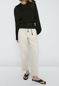 Zwarte gebreide trui met een losse pasvorm en ribbelstructuur, gecombineerd met lichtbeige rechte broeken en zwarte slippers.