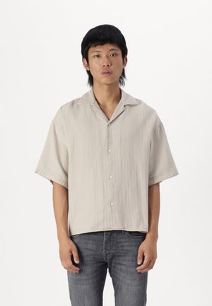 Chemise à manches courtes, beige, avec des boutons, en tissu texturé, col rabattu et coupe décontractée. Associée à un jean en denim foncé.