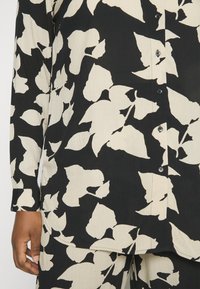 Camicia nera e crema con bottoni, maniche lunghe, caratterizzata da un grande motivo floreale e tessuto testurizzato. I bottoni sul pattino sono visibili.