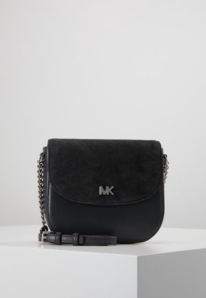 Sac bandoulière en cuir noir et daim avec sangle chaîne argentée et logo "MK" argenté sur le rabat avant, posé sur une surface d'exposition blanche.