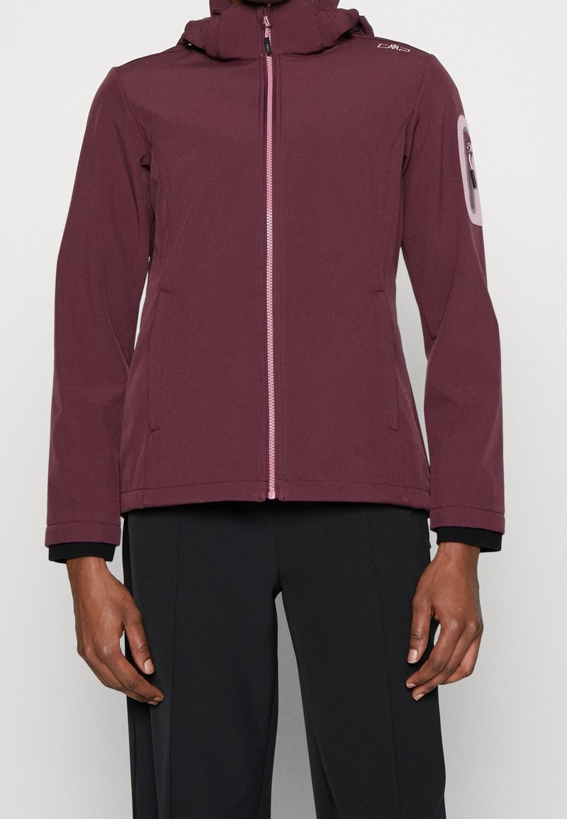 Personne portant une veste zippée bordeaux avec capuche et un pantalon noir, debout devant un fond blanc uni.