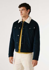 Giacca in velluto a coste blu navy con un morbido colletto in shearling bianco, bottoni frontali, tasche sul petto e un design corto. Indossata sopra un maglione giallo.