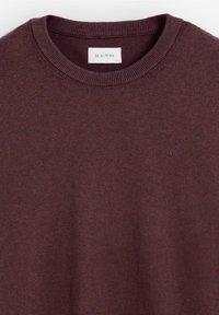 Sweater de punto burdeos con cuello redondo y puños acanalados. Presenta un pequeño logo bordado en el frontal. Textura suave, diseño casual.