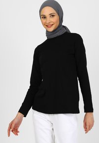 Sort langærmet skjorte lavet af blødt materiale, med en crew neck design. Modellen bærer et gråt hijab og hvide bukser.