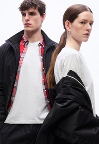Νέος άνδρας με κόκκινο πουκάμισο καρό σε στρώσεις και μαύρο μπουφάν στέκεται δίπλα σε γυναίκα με μακριά μαλλιά που φοράει λευκό πουκάμισο και μαύρο μπουφάν περασμένο πάνω από τους ώμους.