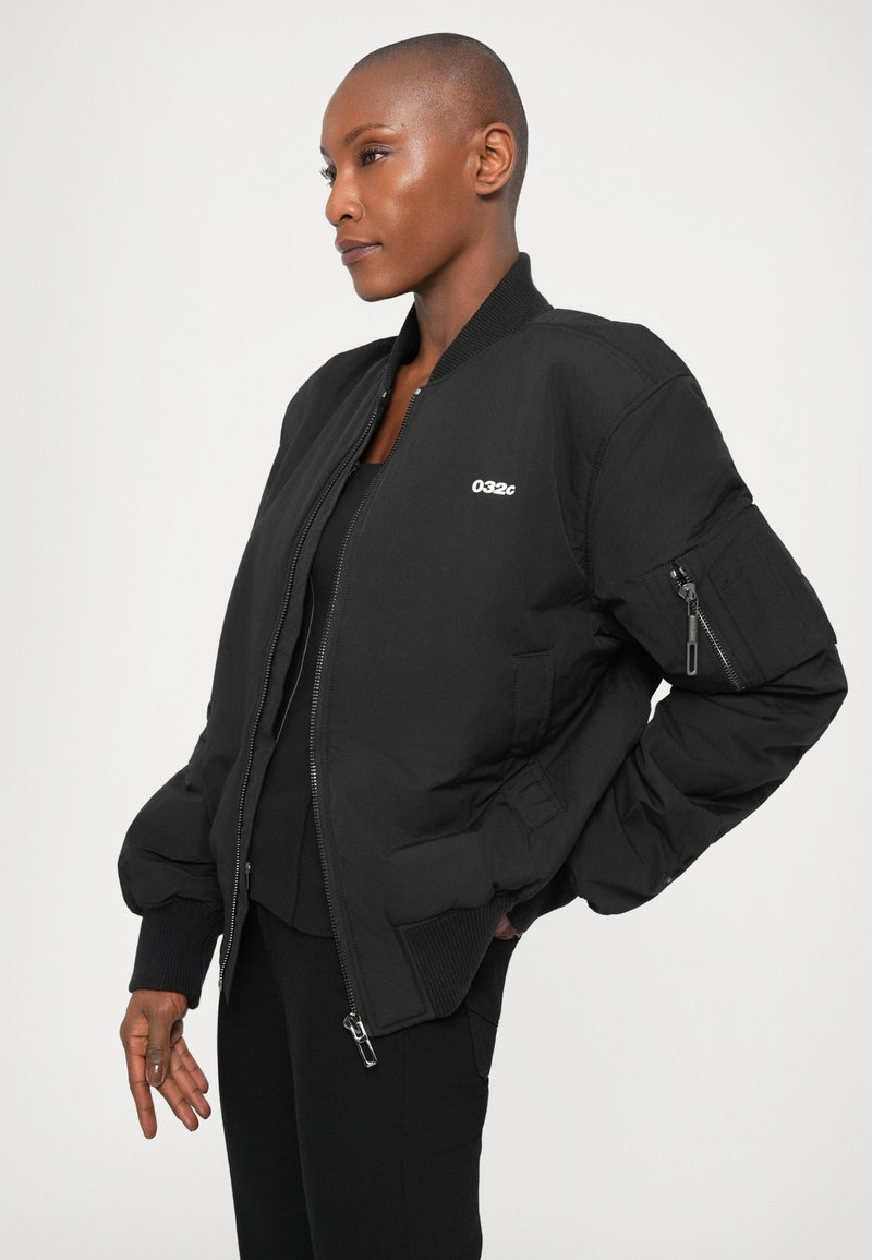 Person with shaved head wearing black bomber jacket with "032c" logo, zipped partially, standing against plain light background.