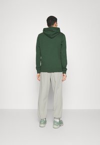 Mand vender væk iført en mørkegrøn hættejakke, lysegrå løse bukser, hvide sokker og grønne sneakers mod en ensartet lys baggrund.