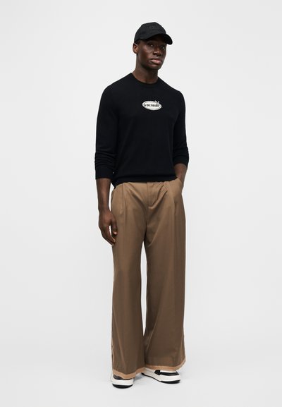 Black sweater with logo, khaki wide-leg trousers, and black cap. Shoes are white with black and gray accents. Smooth fabric texture.