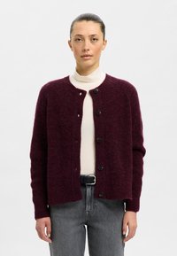 Bordo megzta kardigano su apvalia iškirpte, sagų užsegimu ir ilgomis rankovėmis, derantis su šviesiu golfu ir pilkais džinsais.