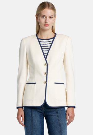 Ivory blazer featuring a notched lapel, gold buttons, navy trim, and two front pockets, layered over a striped top and denim pants.