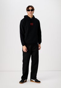 Melna kapuce ar sarkano logo, apvienota ar melnajām biksēm un melnajām sporta kurpēm ar brūnām zolēm. Aksesuāri ietver tumšas saulesbrilles.